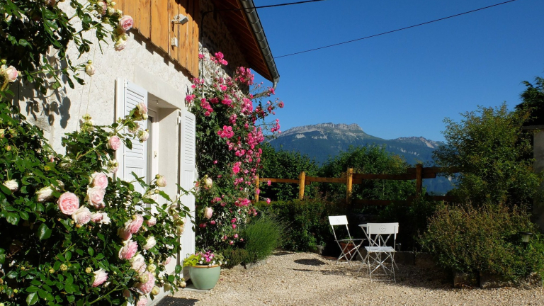 La faade d'une maison abrite baigne dans un soleil radieux. La porte d'entre, cerne de rosiers, ouvre sur une alle o se trouvent une table et deux chaises positionnes prs d'arbustes, orientes vers une montagne et son ciel bleu.