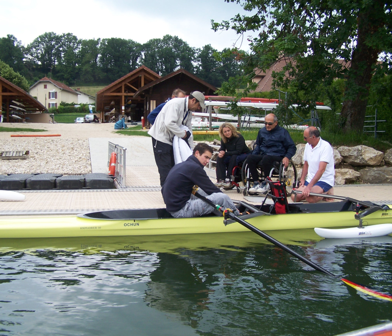 Depuis un quai d'embarquement, une personne en fauteuil roulant, encadre par des moniteurs et ses accompagnateurs, s'apprte  embarquer  bord d'un aviron adapt pour l'accueillir en pratique double.